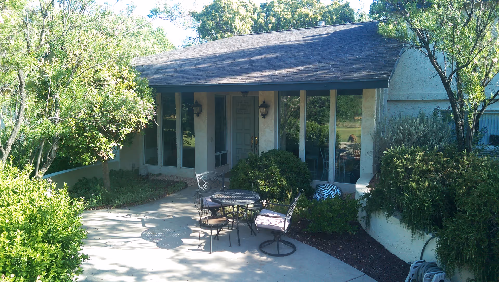 Front patio of a single-story assisted living building with a round table and chairs, large windows, and surrounding shrubs and trees.