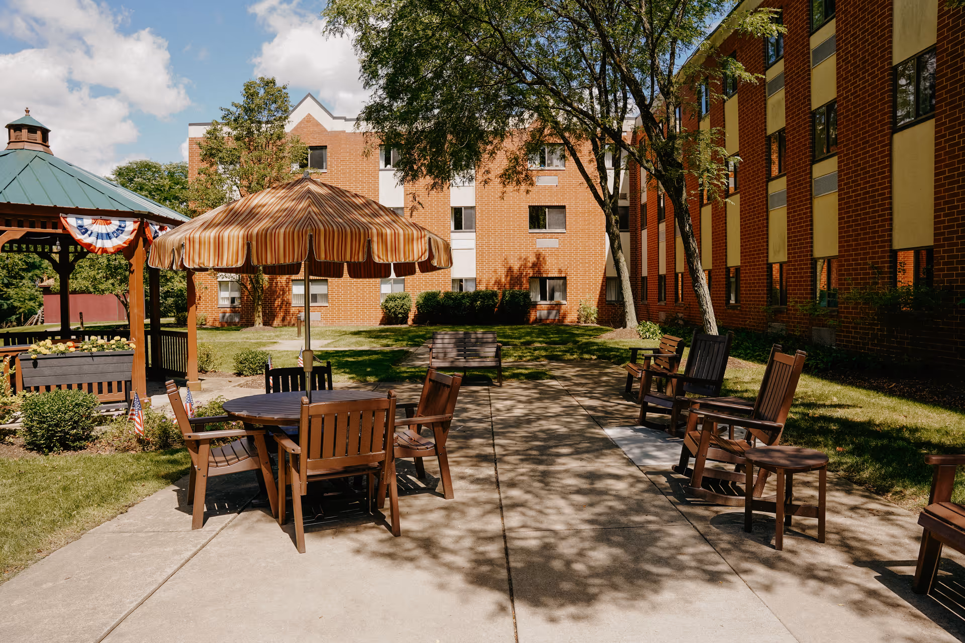 Outdoor courtyard area with wooden tables and chairs, one table shaded by a striped umbrella. There is a gazebo decorated with patriotic bunting on the left side, surrounded by green grass, bushes, and trees. In the background, there are two brick buildings with multiple windows under a partly cloudy sky.