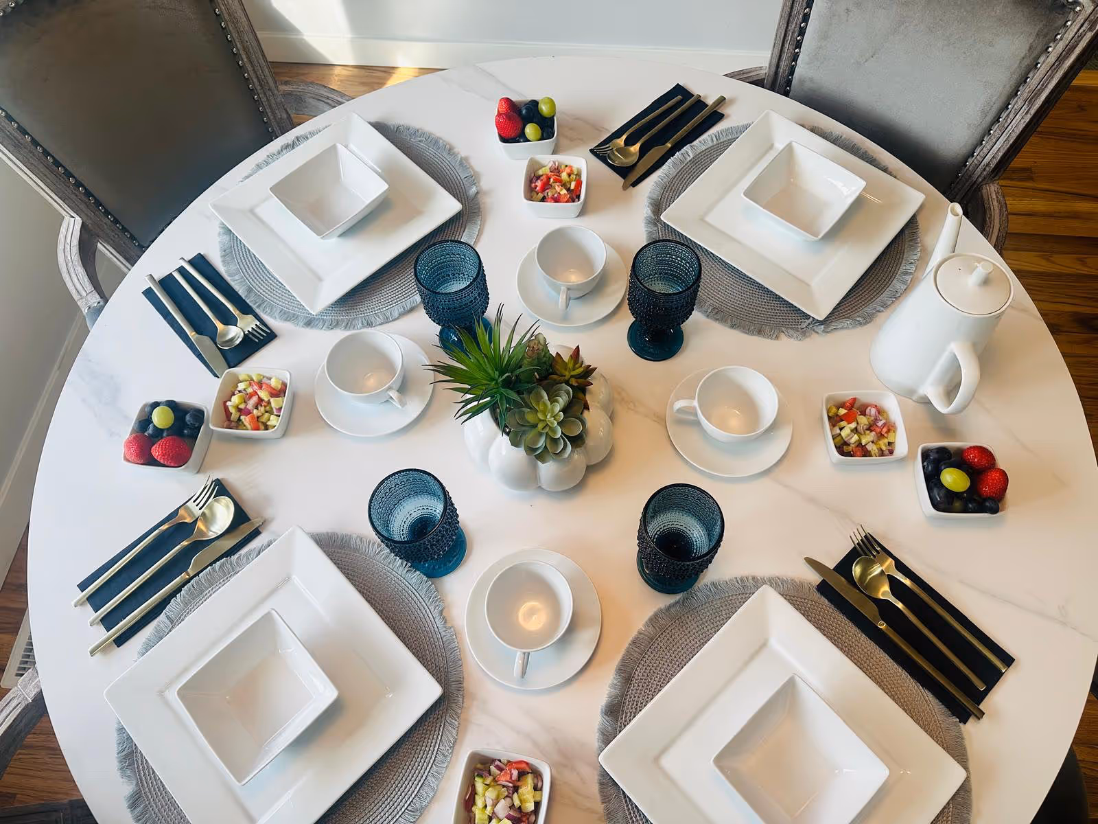 Round marble dining table set for four with plates, cups, cutlery, and a succulent centerpiece.