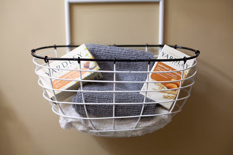 White wire bathroom basket with towel and Yardley boxes mounted on beige wall.