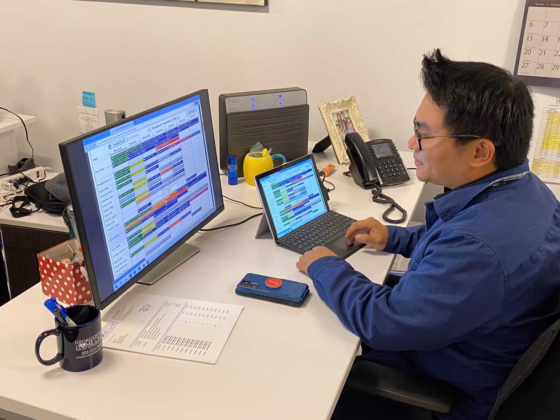 Staff member works at a desk with laptop and large monitor in an office space.
