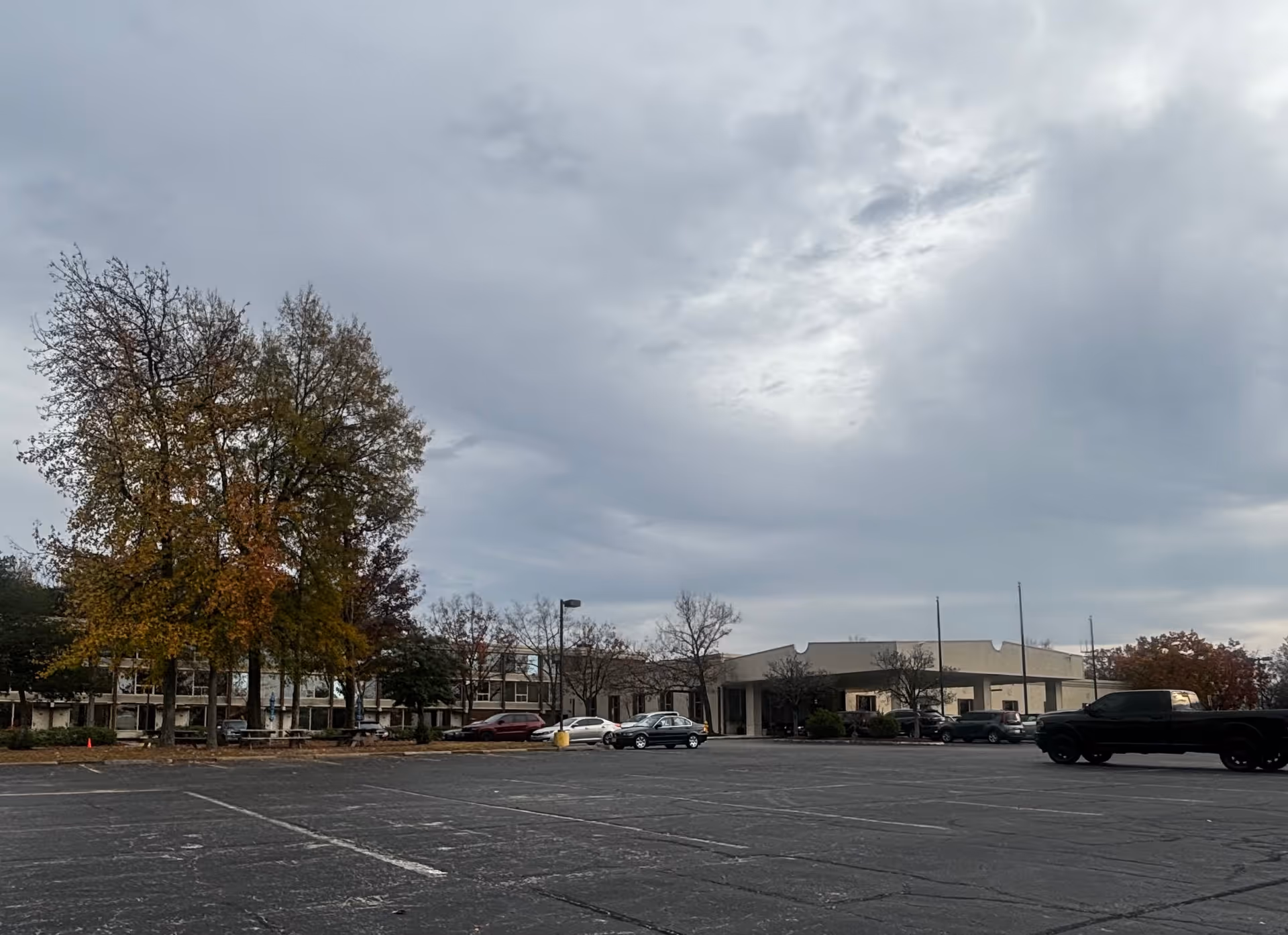 The front of a low-rise senior community building with a mostly empty parking lot, autumn trees, and an overcast sky.
