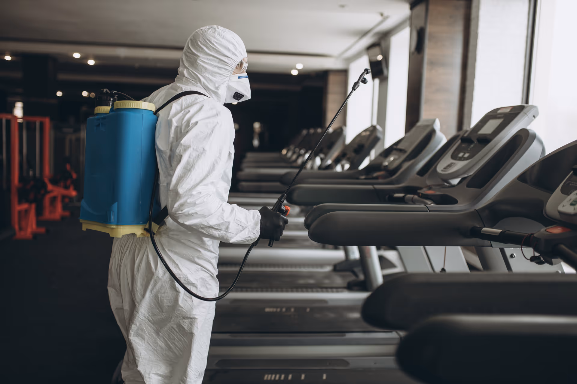 Person in full protective suit disinfects treadmills in a dim fitness center at a senior facility.