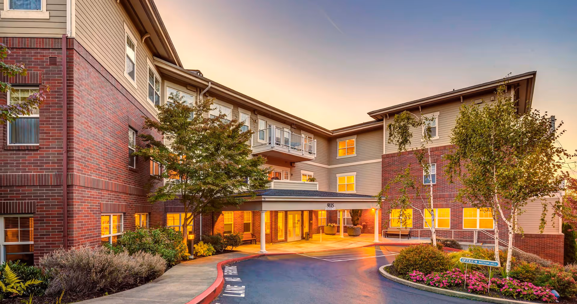 Exterior view of a senior living facility building at sunset with warm lights glowing from the windows. The building features a combination of red brick and beige siding, with a covered entrance supported by white columns. There are trees, shrubs, and flower beds surrounding the driveway and entrance area.