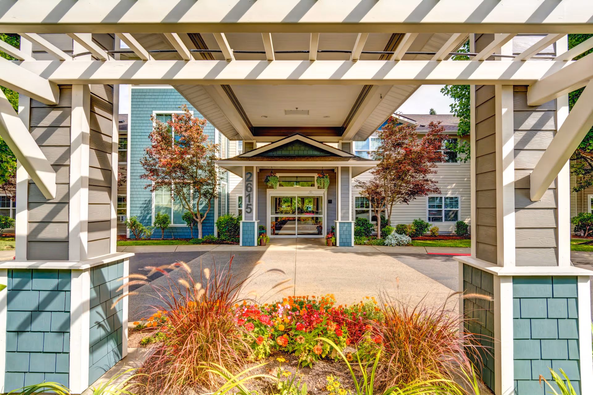 Front entrance of Lone Oak Assisted Living facility with a covered driveway, decorative wooden beams, colorful flower beds, and trees on either side of the entrance. The building has light blue and beige siding with large windows.