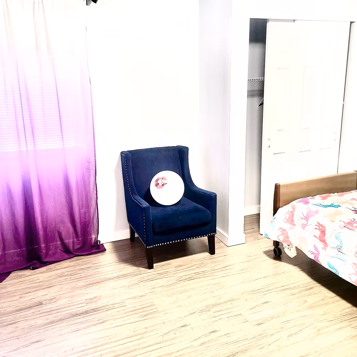 Blue upholstered chair beside a bed in a bright bedroom with a purple curtain.