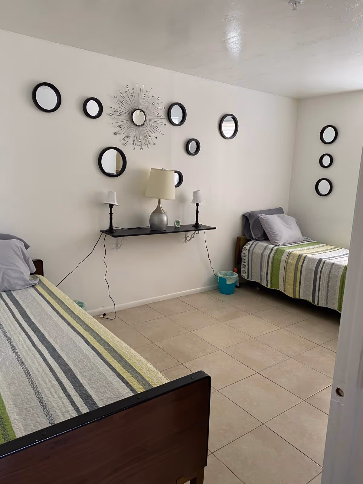 A shared dorm-style bedroom features two single beds, tiled floor, and wall circle mirrors.