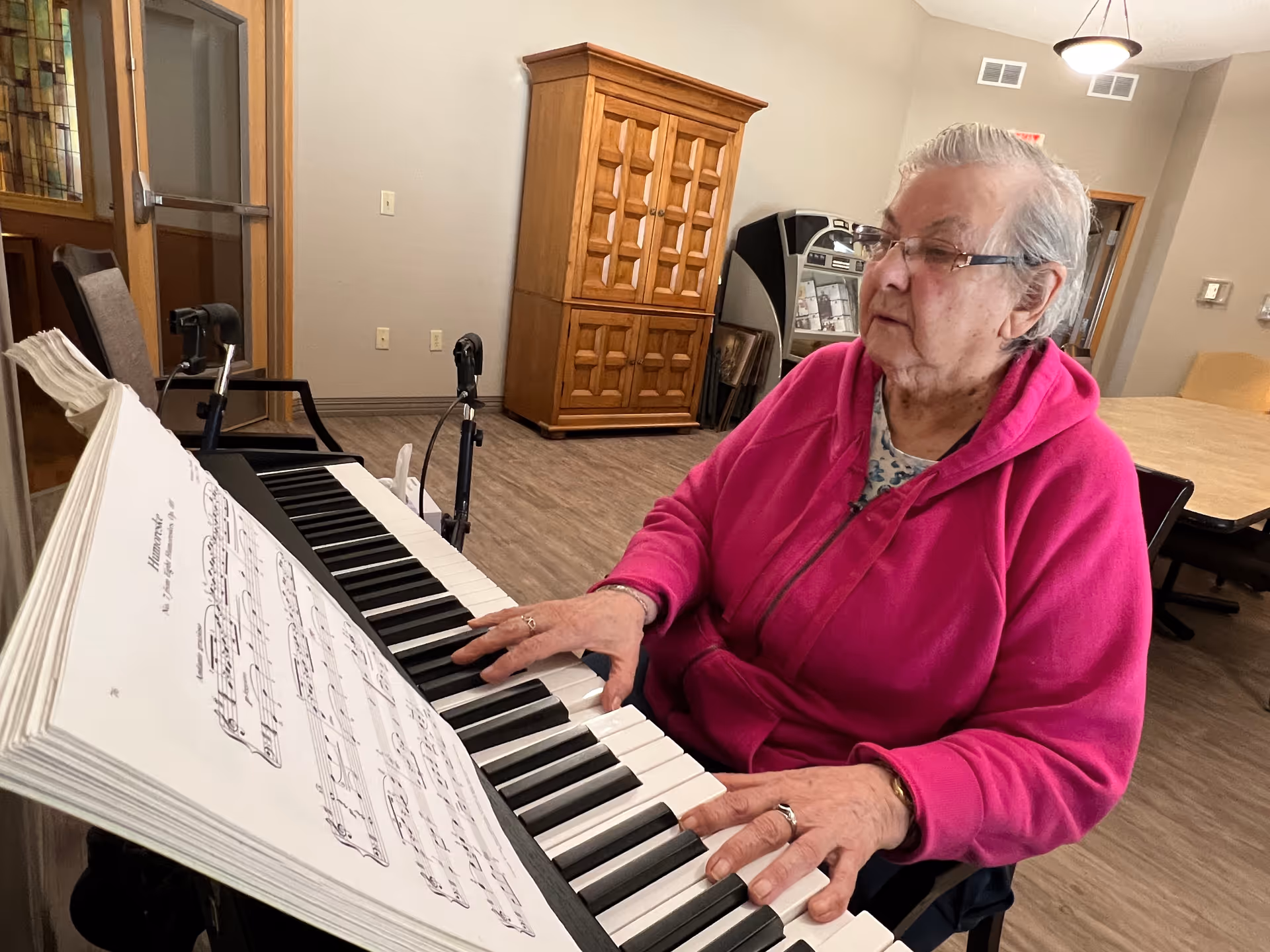 An elderly woman wearing a bright pink hoodie and glasses is playing a keyboard piano with sheet music in front of her. She is seated in a room with wooden flooring, a wooden cabinet, and a table with chairs in the background.