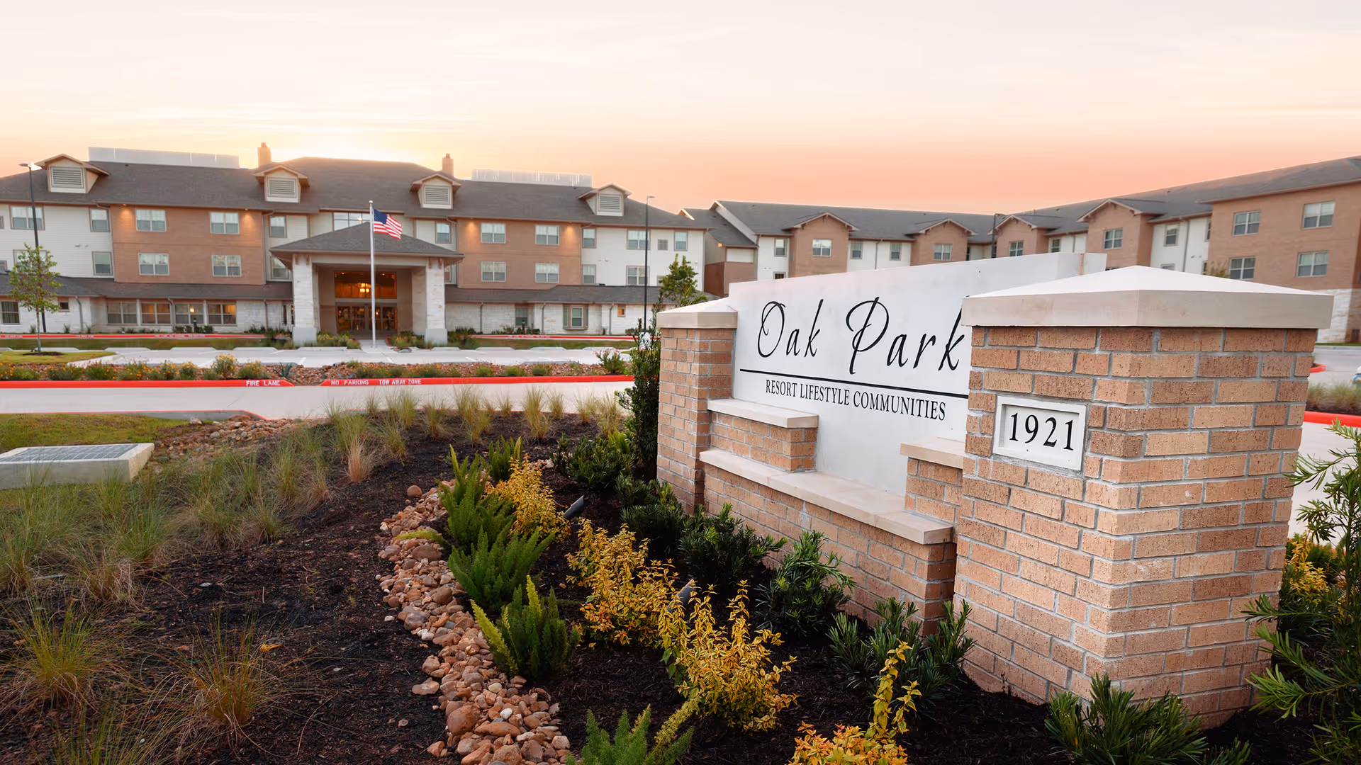Exterior view of Oak Park Retirement Resort at sunset, showing a large multi-story building with multiple windows and a central entrance with an American flag. In the foreground, there is a landscaped area with plants and a brick sign displaying the name 'Oak Park Resort Lifestyle Communities' and the number '1921'.