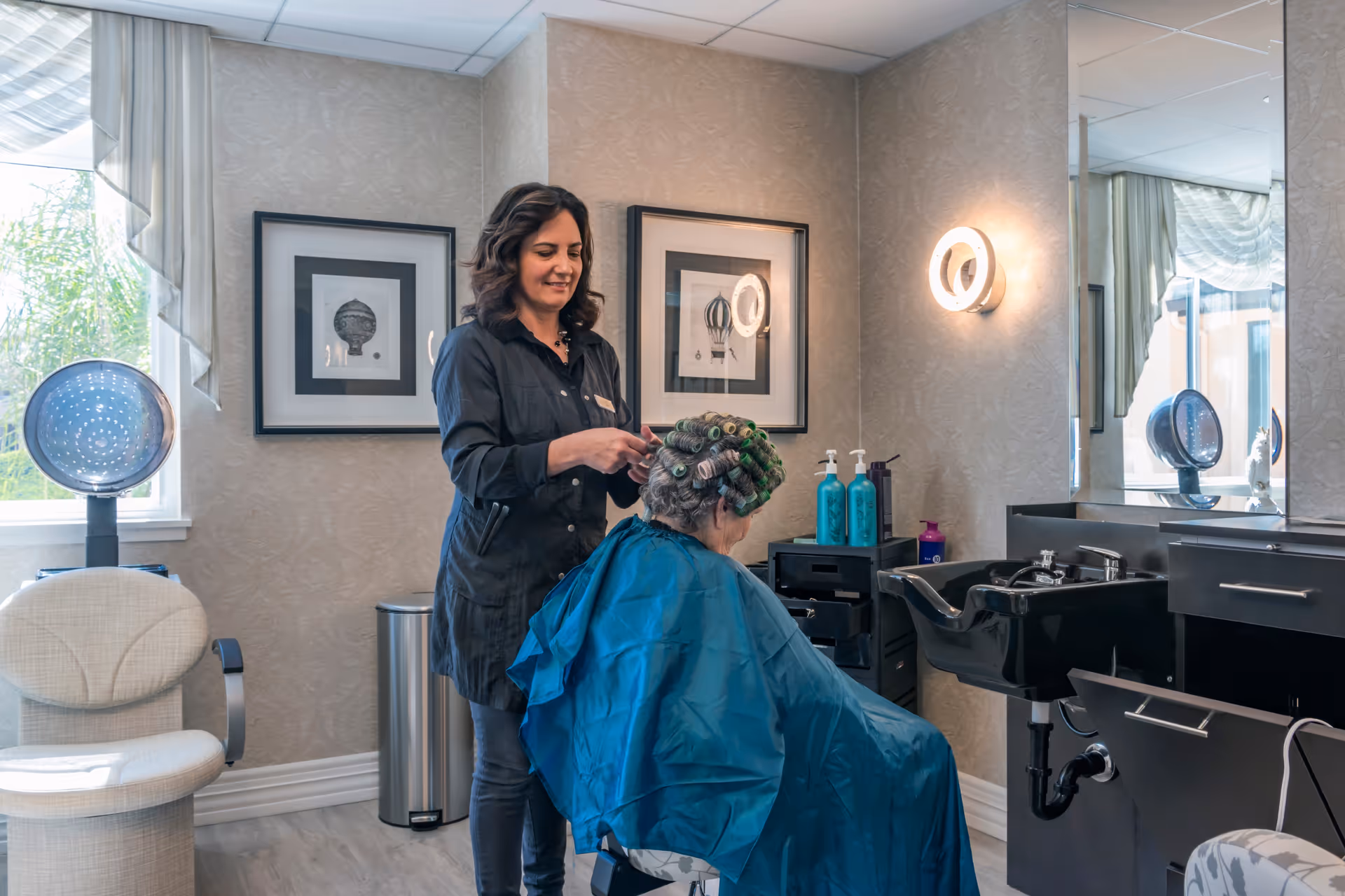 A hairstylist is working on an elderly woman's hair, who is seated with hair rollers and a blue cape in a salon area. The room has a hair washing sink, salon chairs, framed pictures on the wall, and hair care products on a counter.