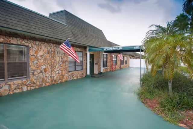 Exterior view of a stone building with a green roof and a covered entrance labeled 'VIP CARE PAVILION 6810'. Two American flags are mounted on the building near the entrance. There is a green paved area in front and some palm trees and greenery on the right side.