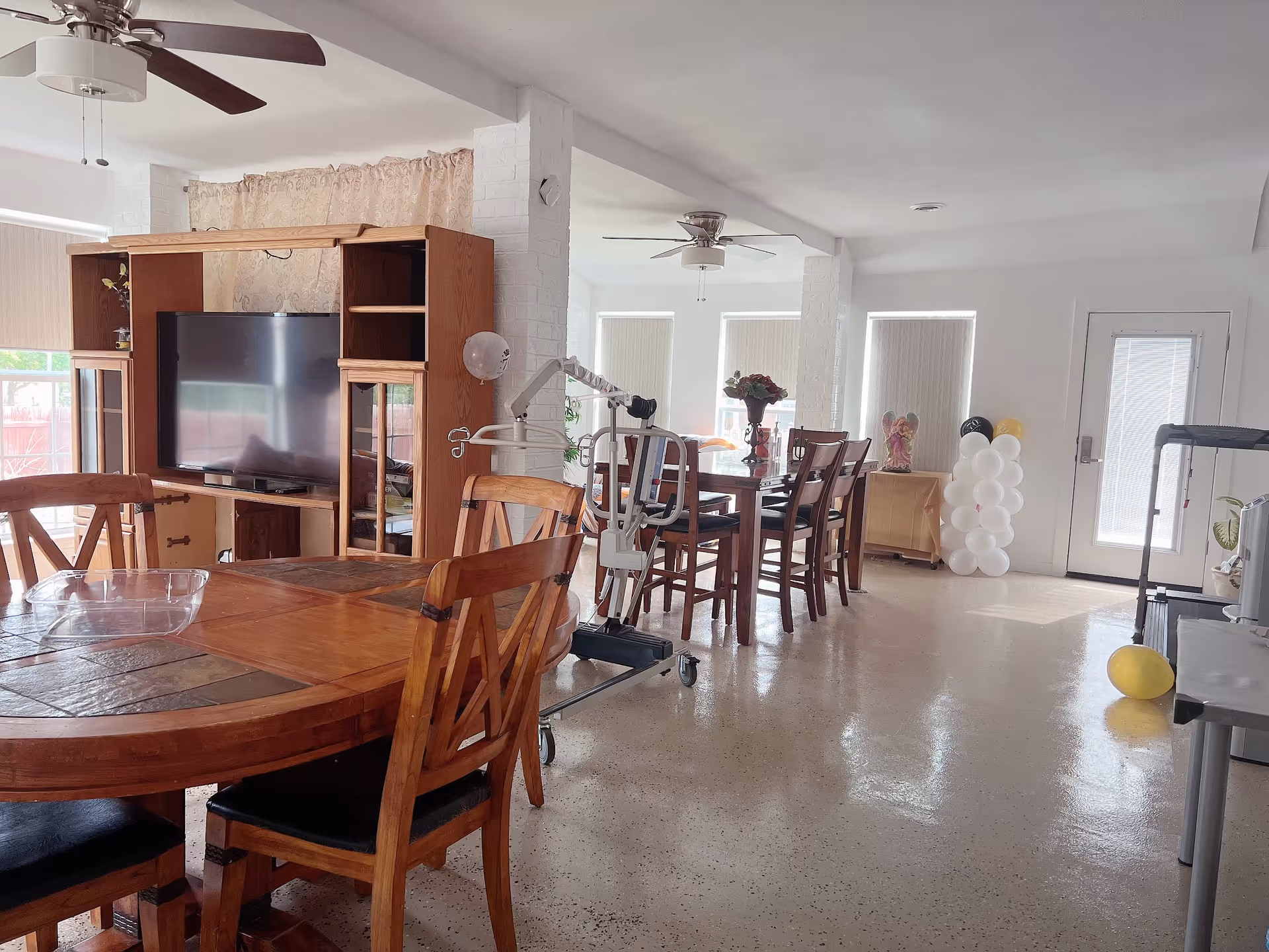 Spacious indoor dining area with wooden tables, chairs, TV cabinet, balloons, and exercise equipment.