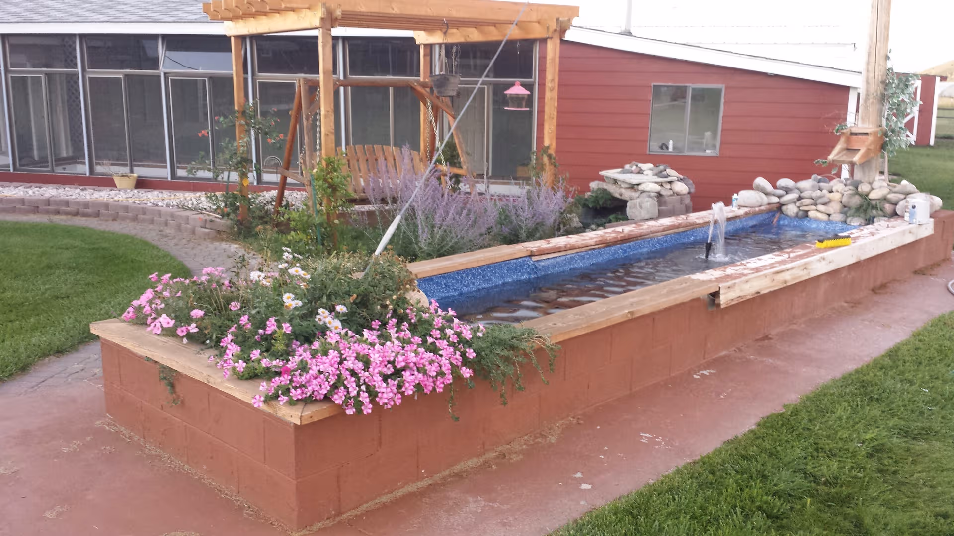 Outdoor courtyard with a long brick planter and fountain, flowers, rocks, and a wooden pergola.