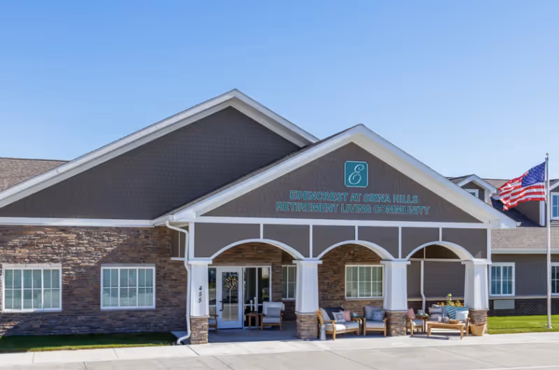 Covered front entrance of the Edencrest at Siena Hills retirement living community showing the building facade, seating area, and an American flag.