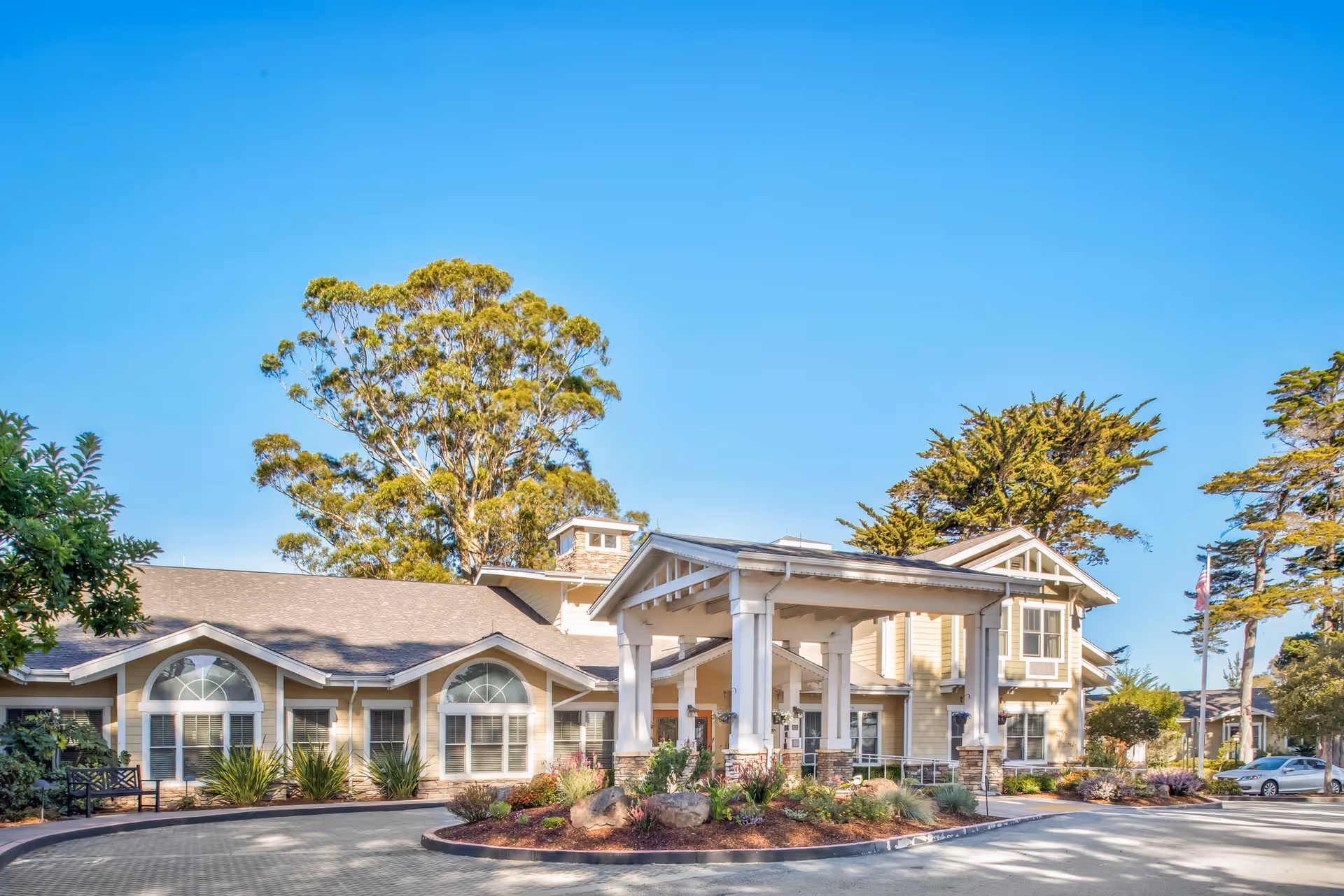 Exterior view of a senior living facility building with a covered entrance, surrounded by trees and landscaping under a clear blue sky.