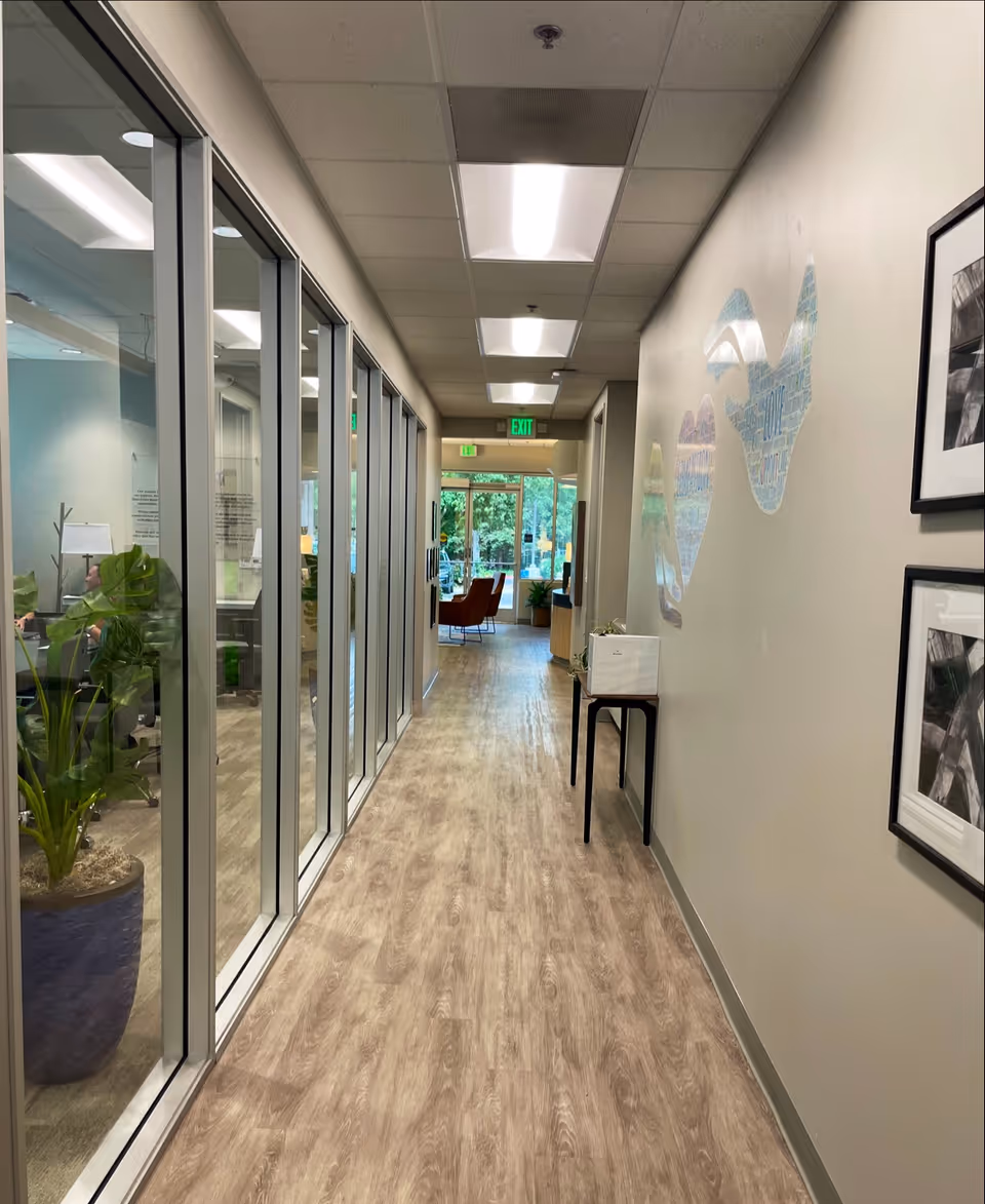 Interior hallway of a senior facility with glass-walled rooms, plants, and exit signs.