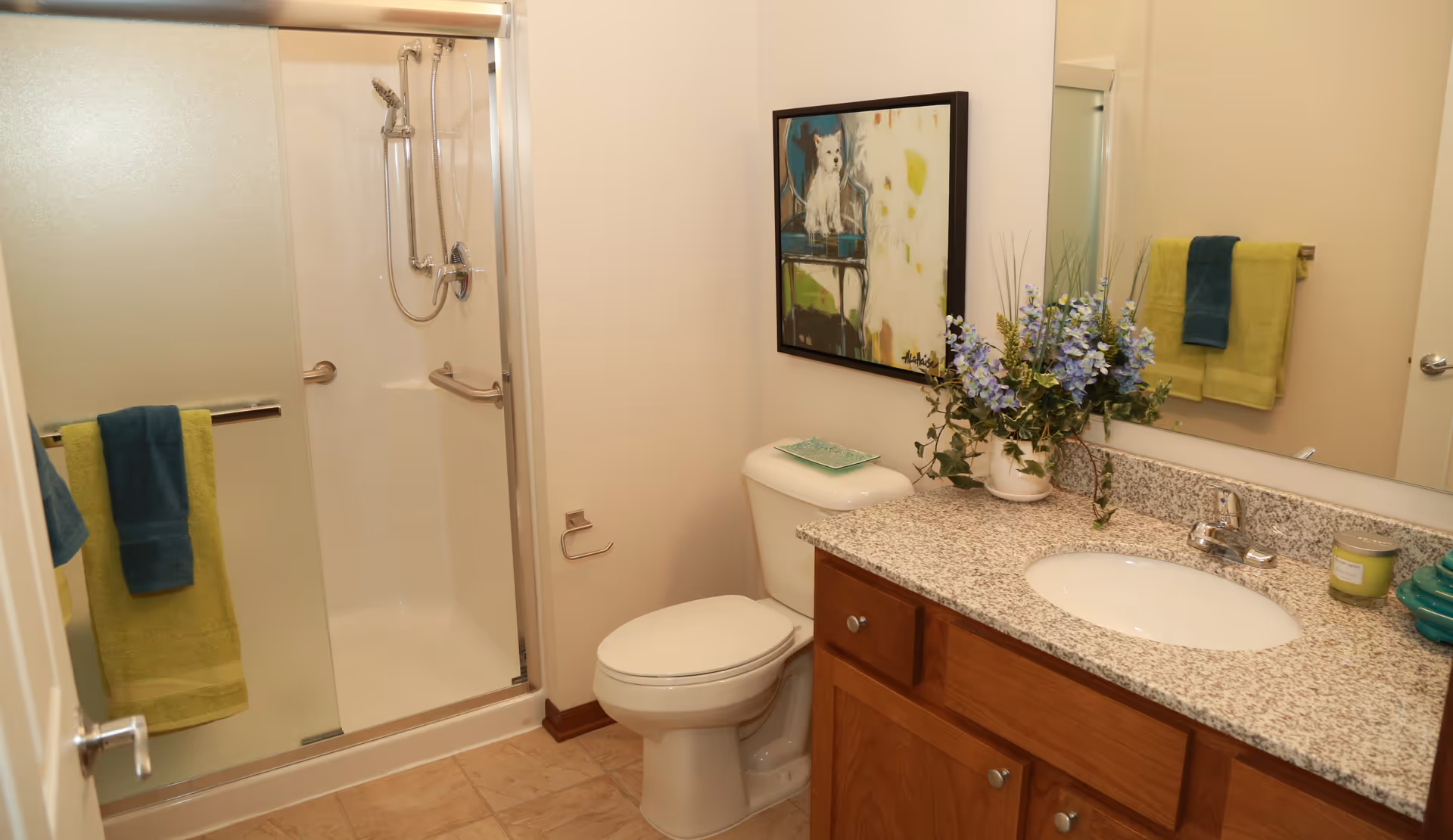 Bathroom with a glass-enclosed shower, toilet, and a granite-topped vanity with sink, towels, and a floral arrangement.