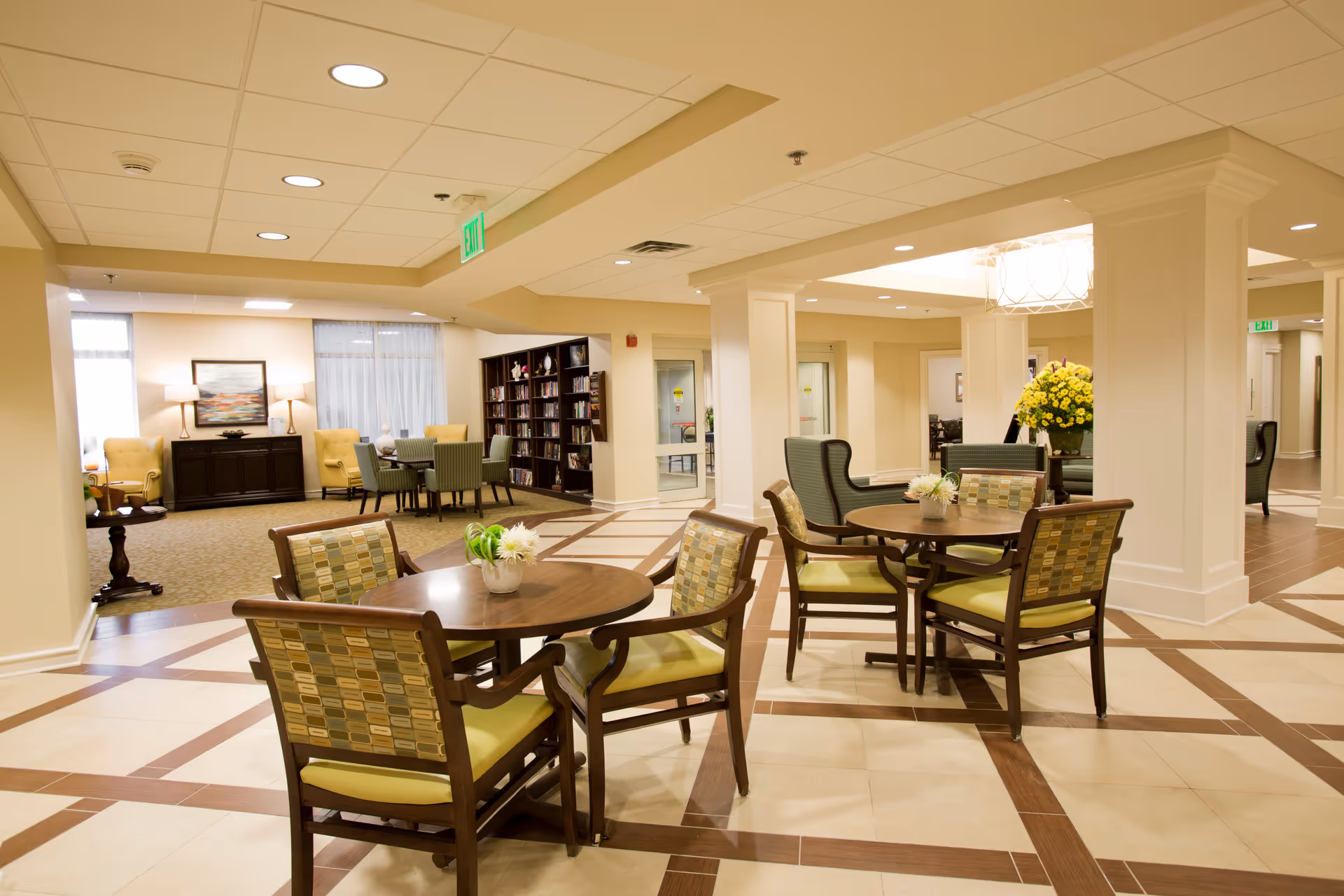 A spacious and well-lit common area in a senior living facility with round wooden tables and cushioned chairs arranged for seating. The room features tiled flooring with a decorative pattern, white pillars, and a bookshelf filled with books in the background. There are also comfortable armchairs and a sideboard with lamps and artwork on the far side of the room.