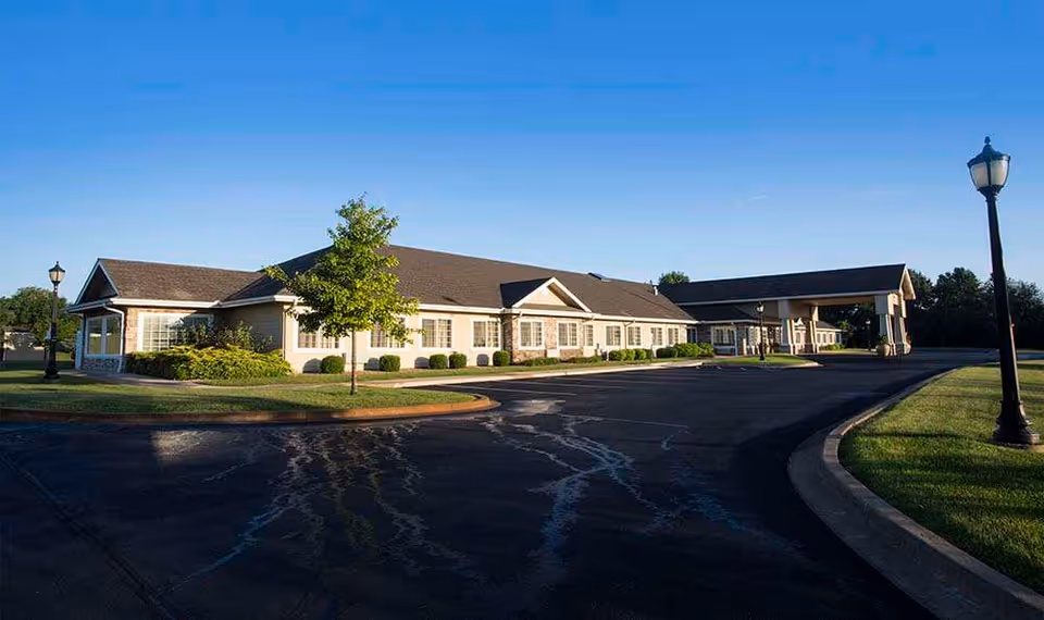 Exterior view of a single-story building with a covered entrance, surrounded by a paved driveway, green grass, small bushes, and a few trees under a clear blue sky.