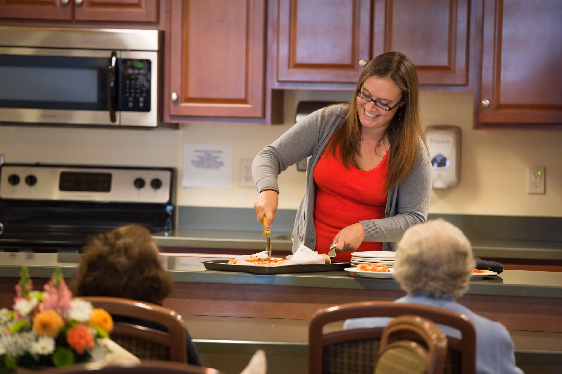 A woman wearing glasses and a red shirt with a gray cardigan is smiling while cutting a pizza on a tray in a kitchen area. Two elderly people are seated in front of her, watching. The kitchen has wooden cabinets, a microwave, and a stove.