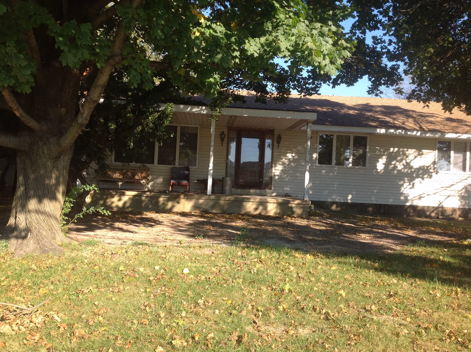 Front exterior view of a single-story building with a covered porch, two chairs, and large trees providing shade. The building has light-colored siding and multiple windows.