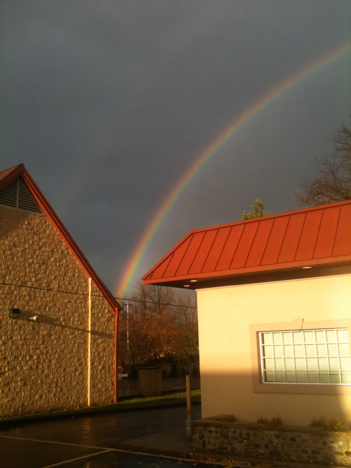 Two tan brick buildings with red roofs in a parking lot beneath a double rainbow.