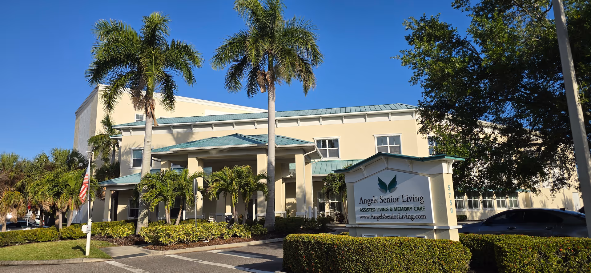 Beige senior living building with palm trees, a sign, and manicured shrubs at the entrance.