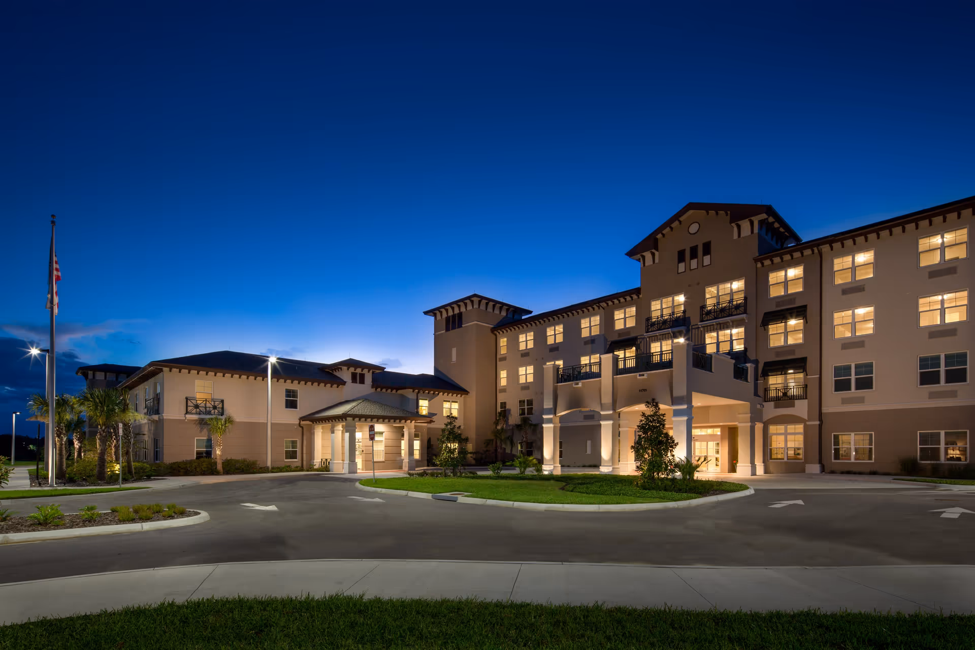 Front exterior of a well-lit multi-story senior living building at dusk with a circular driveway and flagpole.