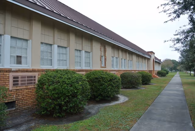 Exterior view of a single-story building with multiple windows and a brown roof, bordered by neatly trimmed bushes and a sidewalk running alongside the building under an overcast sky.