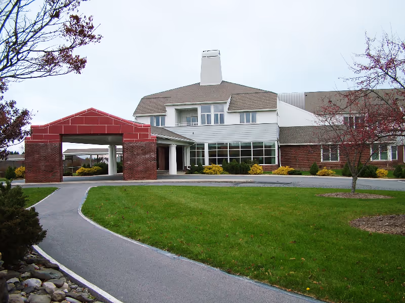 Exterior view of a senior living facility building with a covered entrance supported by brick pillars, a paved driveway, green lawn, and some trees with sparse leaves.