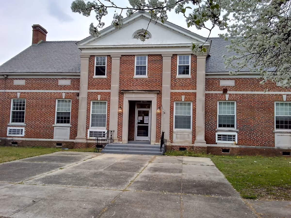 Front facade of a red-brick two-story building with a central columned entrance, steps, and a paved driveway.
