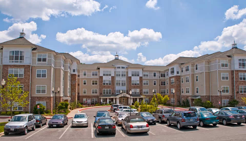 Front exterior view of a large senior living facility building with multiple floors, beige and brick facade, several windows, and a parking lot filled with cars in front under a partly cloudy sky.