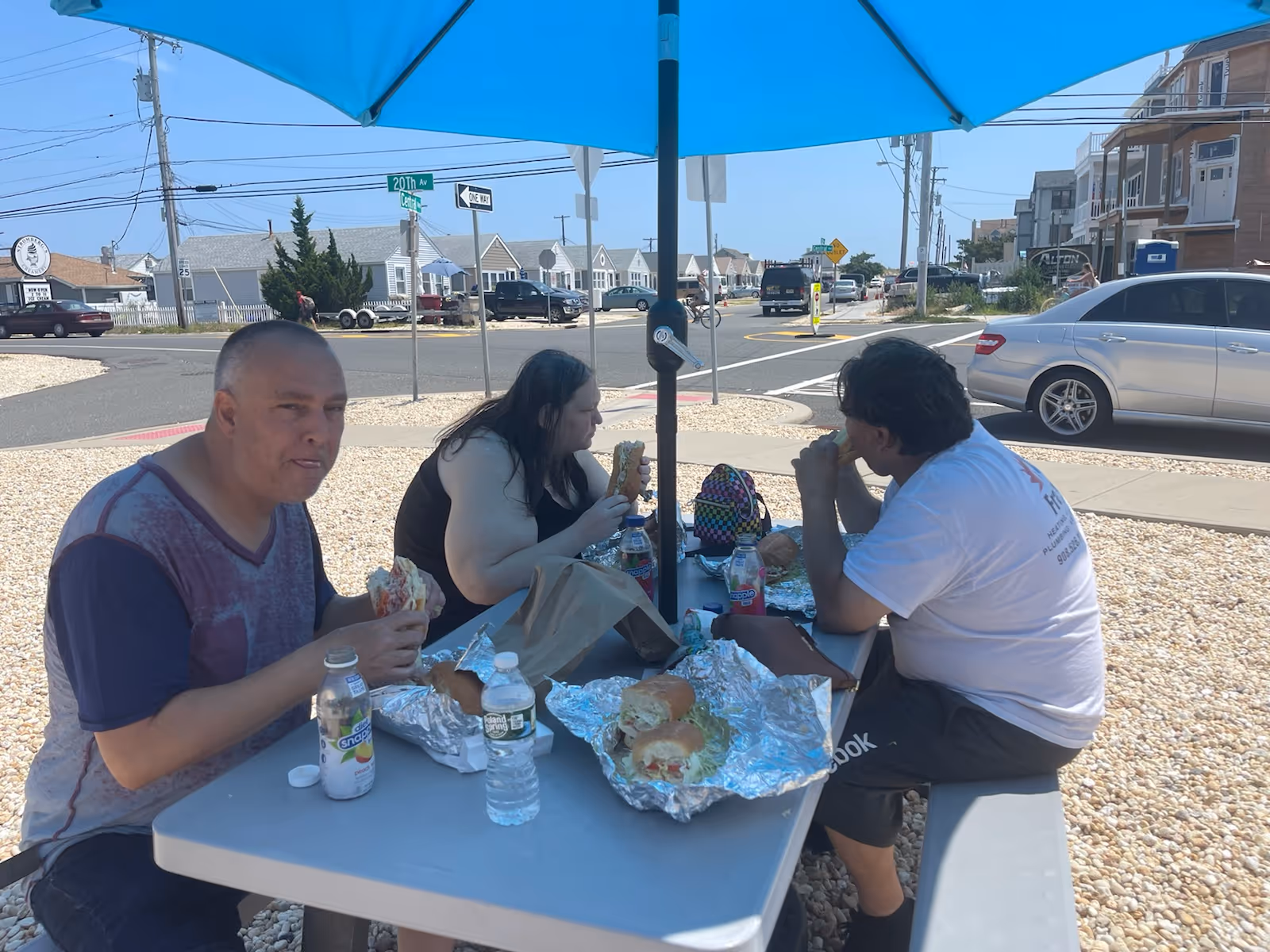 Three people eat sandwiches at a shaded outdoor picnic table under a blue umbrella.