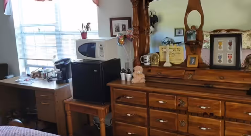 A bedroom with a wooden dresser, mirror, microwave, and a desk near a window.