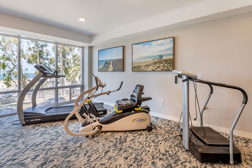 Interior senior living gym with two treadmills and a NuStep bike by windows.