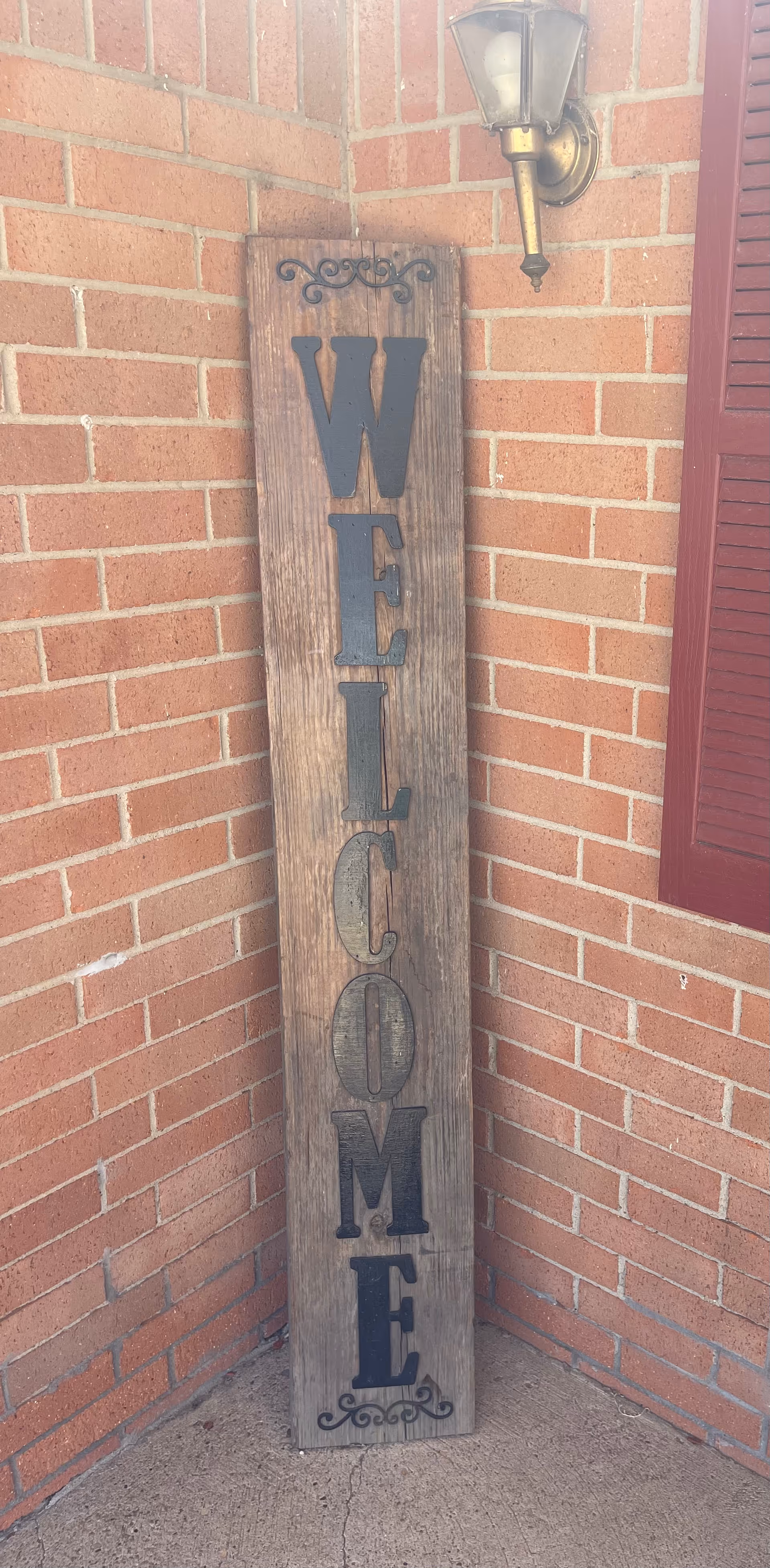 A tall wooden sign leaning against a brick wall with the word 'WELCOME' written vertically in large black letters. Above the sign is a brass outdoor wall lantern, and to the right is a red window shutter.