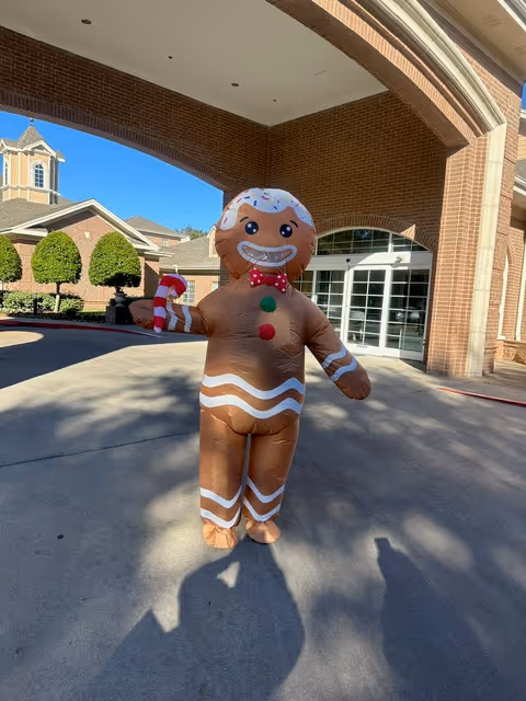An inflatable gingerbread figure stands outside a brick building entrance on a sunny day.