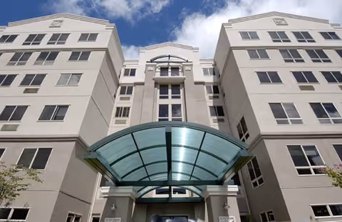 A multi-story senior living building with a curved glass entrance canopy against a blue sky.
