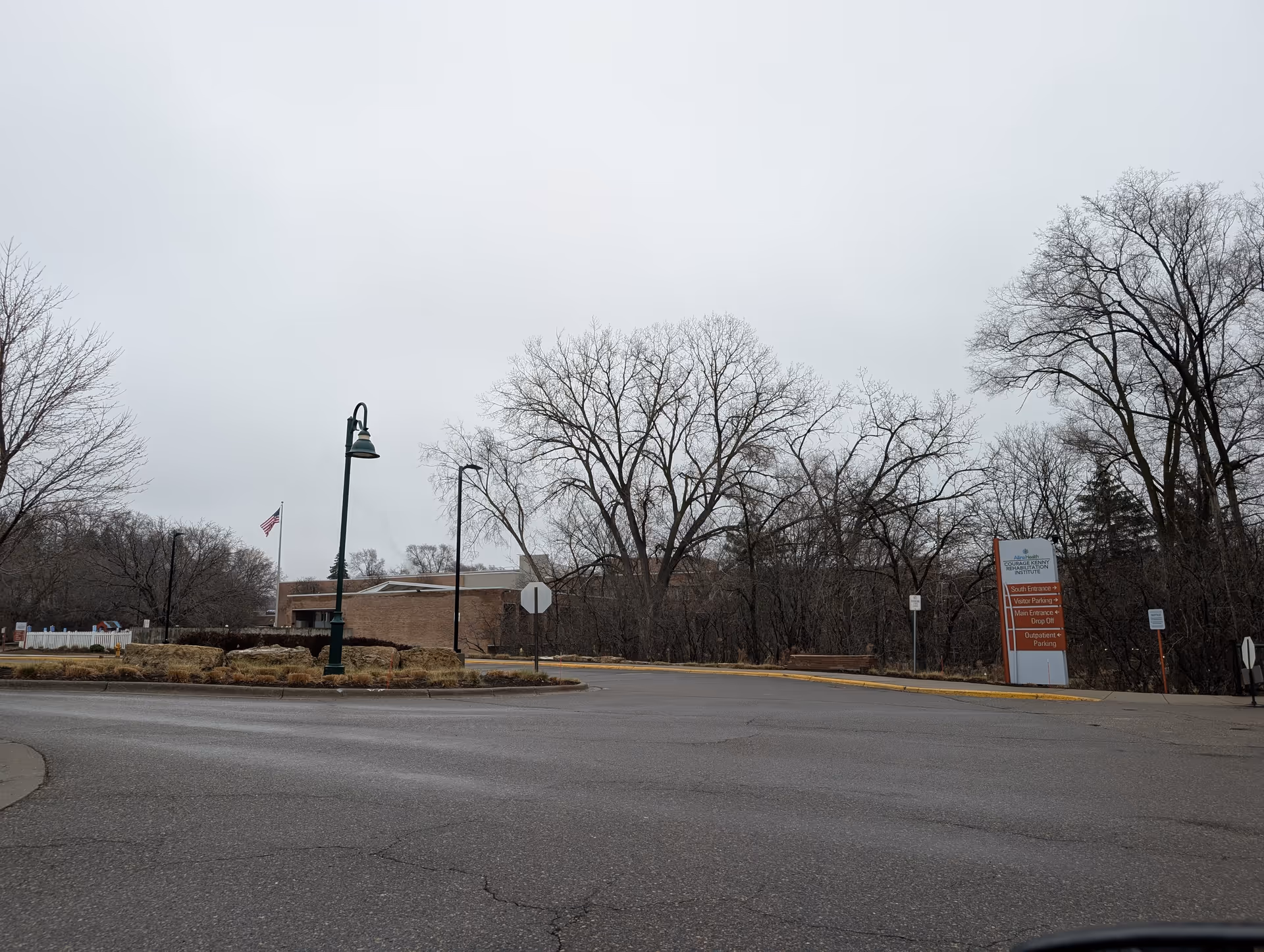 Exterior view of a low brick rehabilitation building and entrance sign beside a circular driveway, leafless trees and an overcast sky.