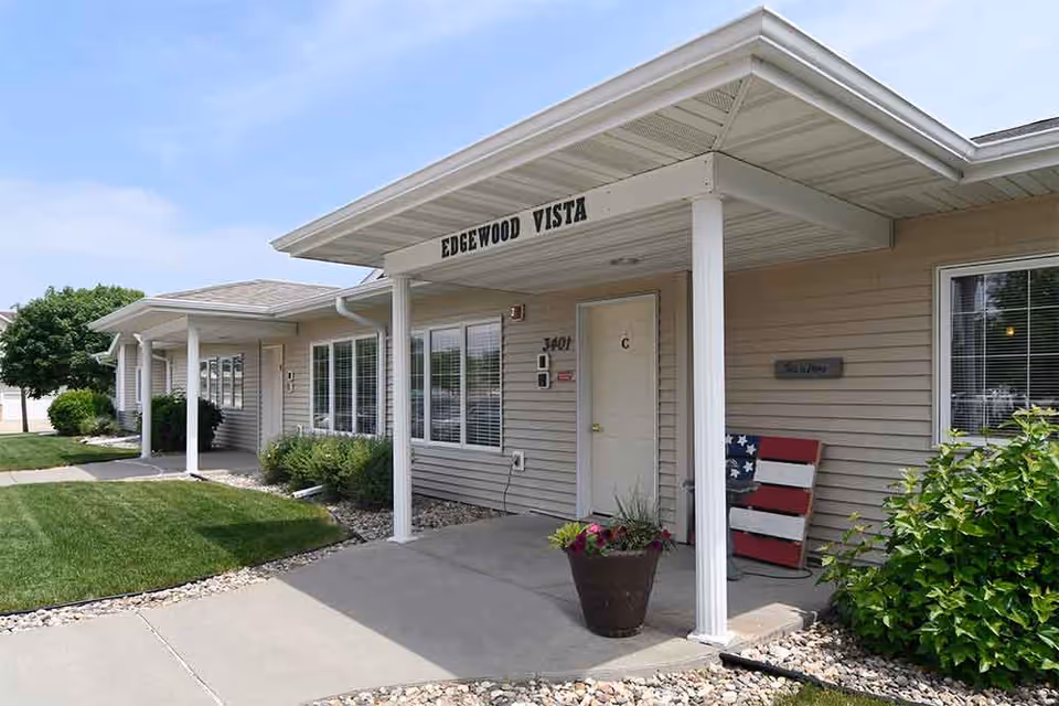 Exterior view of a single-story building with beige siding and white trim. The entrance has a covered porch supported by white columns, with the sign 'EDGEWOOD VISTA' above. There is a potted plant with flowers near the door, a decorative American flag-themed chair, and well-maintained landscaping with green grass, bushes, and rocks along the walkway.