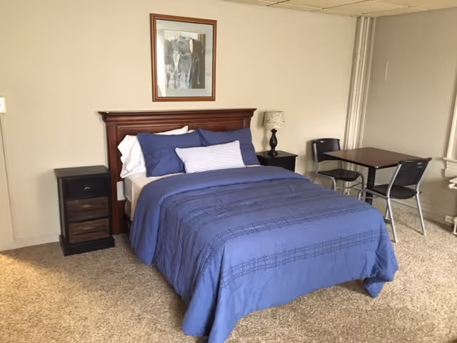 A bedroom with a bed covered in blue bedding and multiple pillows, flanked by two dark wooden nightstands. One nightstand has a lamp with a patterned shade. There is a small table with two chairs near a window on the right side. A framed picture hangs on the wall above the bed.