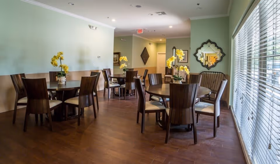Communal dining room with round wooden tables and chairs, yellow flower centerpieces, and large window blinds.