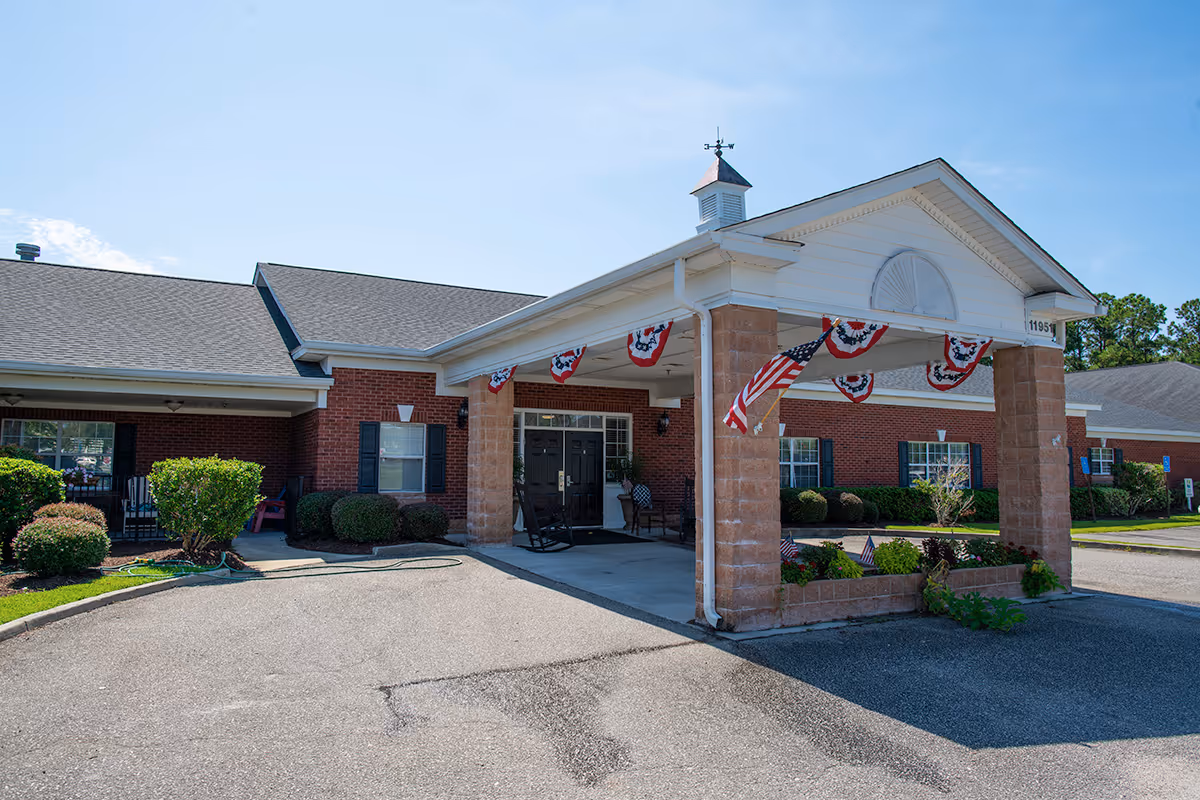 Exterior view of a single-story brick building with a covered entrance supported by brick columns. The entrance area is decorated with patriotic bunting and an American flag. There are bushes and small plants around the building, and the sky is clear and blue.