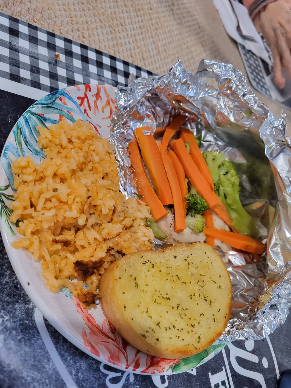 A plate with cooked rice, a piece of garlic bread, and foil containing steamed vegetables including carrots and broccoli, placed on a table with a checkered and textured tablecloth. A person's hand is partially visible in the background.