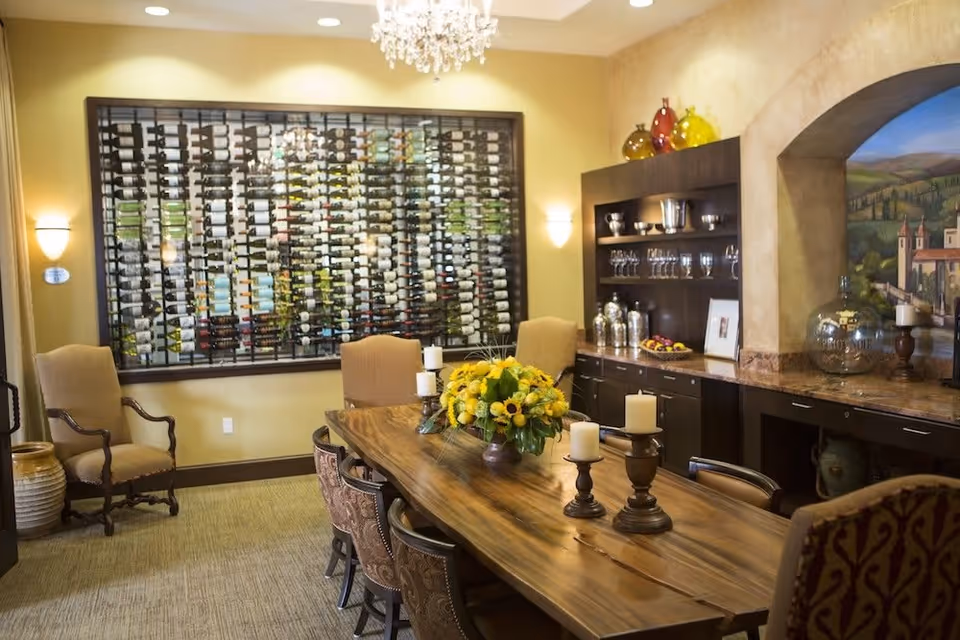 A cozy dining room with a long wooden table surrounded by upholstered chairs. The table is decorated with a floral centerpiece and several candles. On the wall, there is a large wine rack filled with bottles. To the right, there is a built-in cabinet with glassware and decorative items, and a mural depicting a scenic landscape is visible through an arched opening.