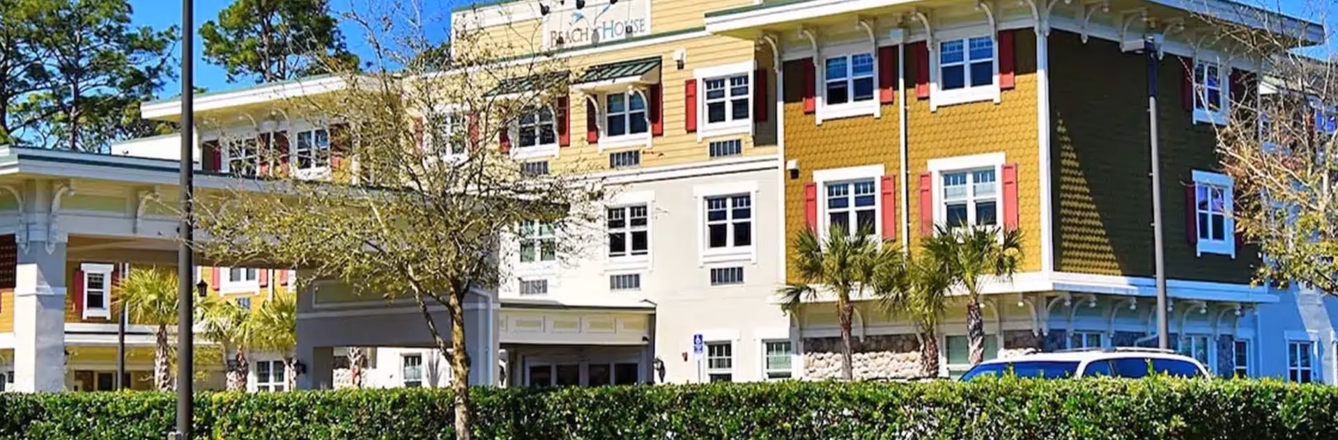 Exterior view of a multi-story assisted living facility named Beach House Assisted Living & Memory Care, featuring a yellow and white facade with red window shutters, palm trees, and a covered entrance with a driveway. There is greenery and a hedge in the foreground under a clear blue sky.