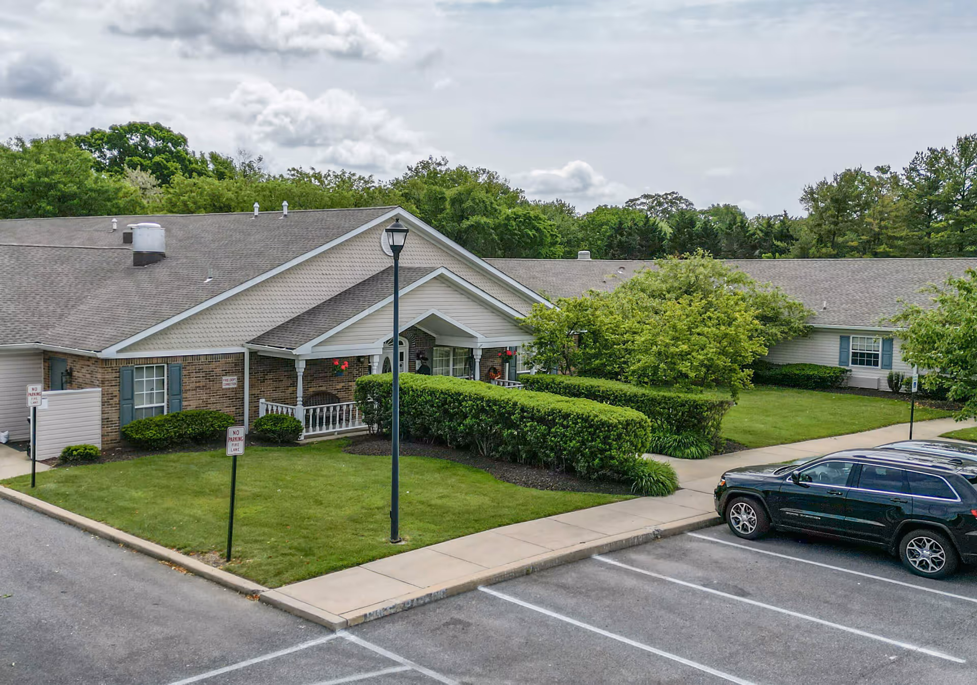 Exterior view of a single-story brick and siding building with a gabled roof, surrounded by green bushes, trees, and a well-maintained lawn. A black SUV is parked in the parking lot adjacent to the building. The sky is partly cloudy.
