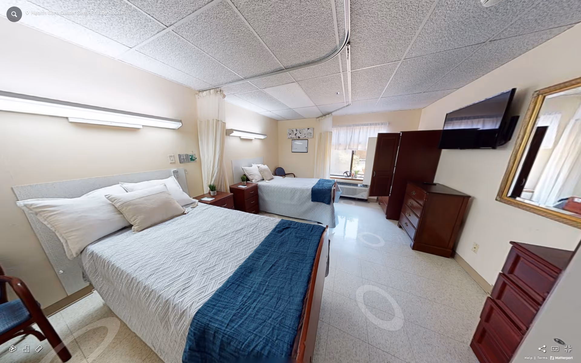 A semi-private room in a senior care center with two beds, each made with white bedding and blue throws. The room has beige walls, a window with sheer curtains, a wall-mounted TV, wooden dressers, a mirror, and overhead fluorescent lighting.