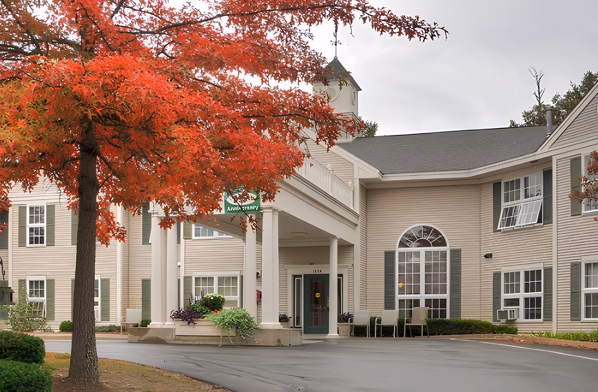 Exterior view of Summercrest Senior Living Community building with beige siding, green shutters, and a covered entrance. A tree with vibrant red autumn leaves is in the foreground.