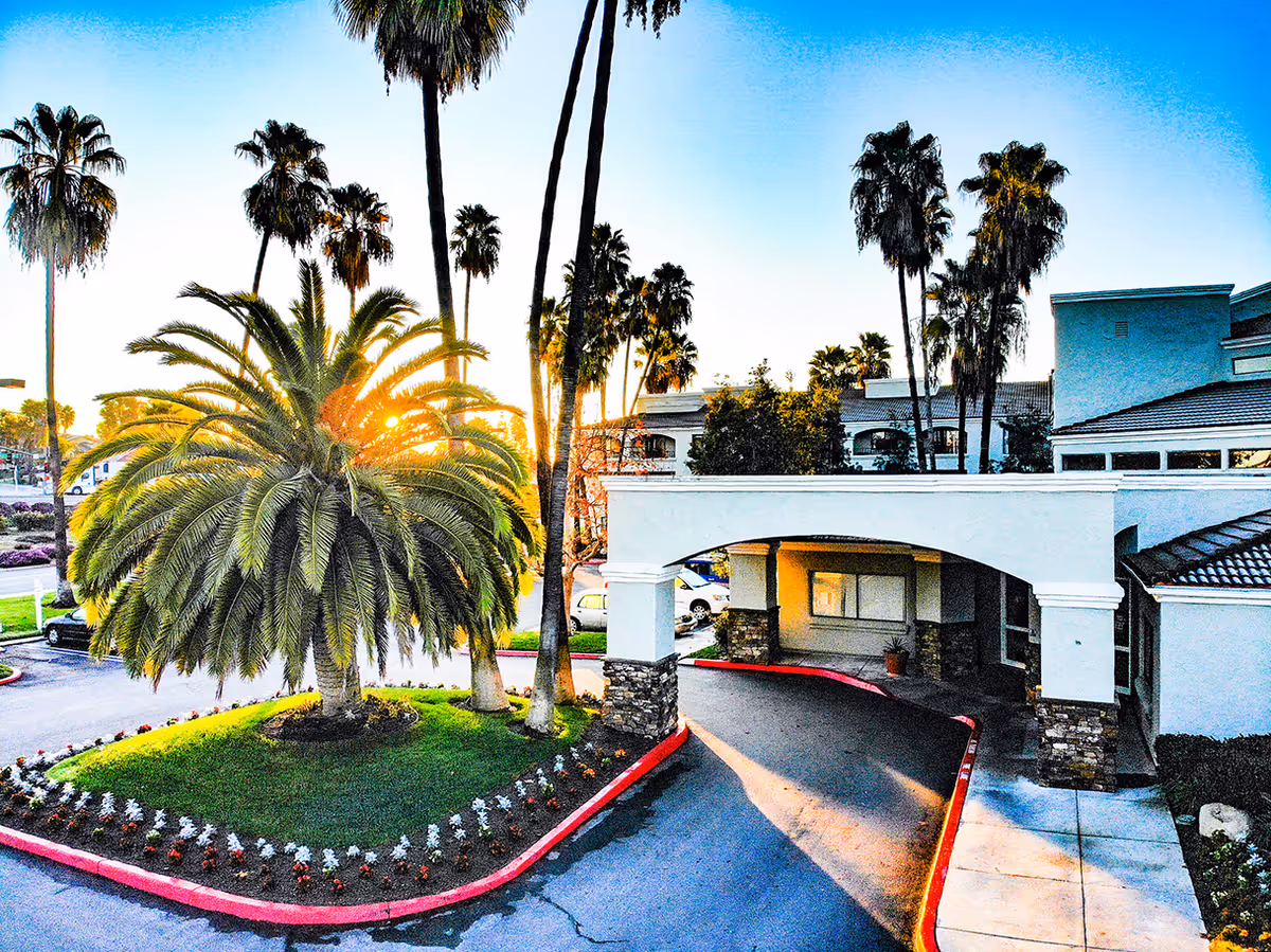 Entrance of Las Villas Del Norte senior living facility with palm trees and landscaped area.